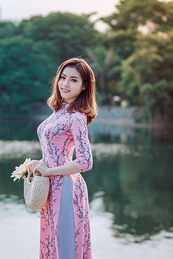 A smiling woman poses gracefully in a pink dress by a serene lakeside, holding a woven basket. HD phone wallpaper