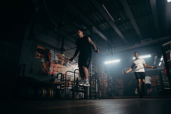 Two men actively skipping jump ropes in a dimly lit gym environment. HD wallpaper