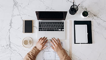 Top view of a stylish home office desk with a laptop, planner, and coffee cup, showing hands on a blueprint. HD wallpaper