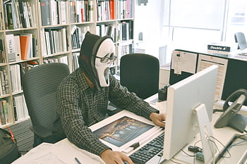 Person wearing a ghost mask working in an office environment filled with books. HD wallpaper