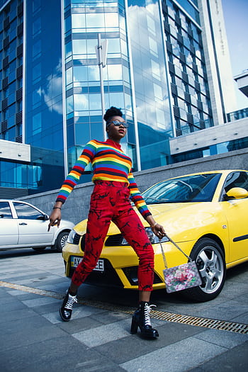 Stylish individual in rainbow top and red pants walking by yellow car in urban area. HD phone wallpaper