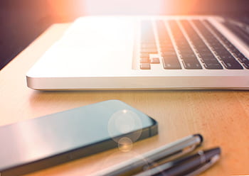 Stylish desk setup featuring a laptop, smart, and pens in natural light. HD wallpaper