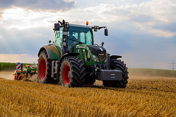 A powerful green tractor plowing a dusty wheat field under a cloudy summer sky in rural Germany. HD wallpaper