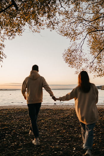 A romantic couple holding hands while walking towards the sunset by the beach, surrounded by autumn scenery. HD phone wallpaper