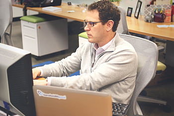Focused man working at a computer in a modern office cubicle, wearing glasses and heads. HD wallpaper