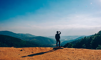 A lone traveler stands on a hilltop, gazing over a vast mountain landscape under a clear blue sky. HD wallpaper