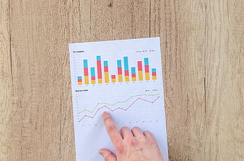 A hand points to colorful business charts and graphs on a paper sheet on a wooden desk. HD wallpaper