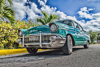 A beautifully restored classic car on a sunny road in Havana, Cuba, with lush foliage and a vibrant sky. HD wallpaper