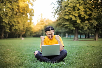 A young man in yellow celebrates success on his laptop while sitting in a sunlit park. HD wallpaper