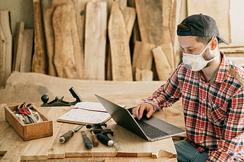 A craftsman wearing a mask, working on a laptop in a woodworking shop. HD wallpaper