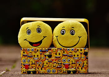 Two yellow smiley plush toys in an emoji-themed box, expressing fun and happiness. HD wallpaper