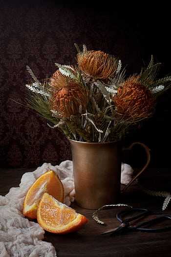 Artistic arrangement of dried flowers in a metallic vase with sliced oranges on a rustic table. HD phone wallpaper