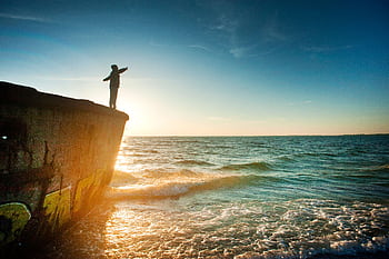 A person stands on a cliff overlooking the ocean at sunset, arms raised in a moment of joy and reflection. HD wallpaper