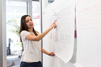 Confident young woman presenting a design on a whiteboard in a modern workspace. HD wallpaper