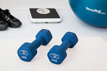 Blue dumbbells placed on a white surface with fitness equipment in the background. HD wallpaper