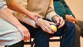 An elderly person receives support from a caregiver, holding hands indoors, showcasing compassion. HD wallpaper