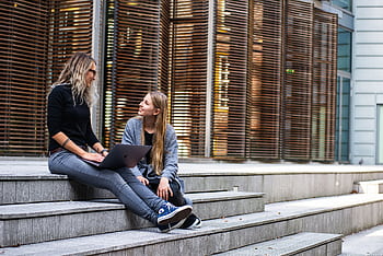 Two women sitting on steps with a laptop, discussing ideas in urban setting. HD wallpaper