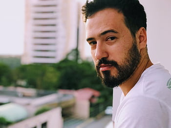 A serious-looking man with a beard stands on a balcony with blurred city buildings in the background. HD wallpaper