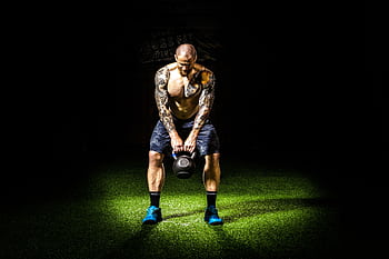 Muscular man with tattoos lifting a kettlebell indoors under a spotlight. Intense fitness and strength training. HD wallpaper