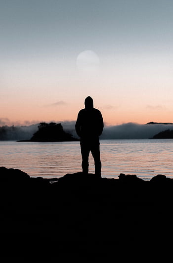 A lone person stands silhouetted against a tranquil sunset over a calm body of water. HD phone wallpaper