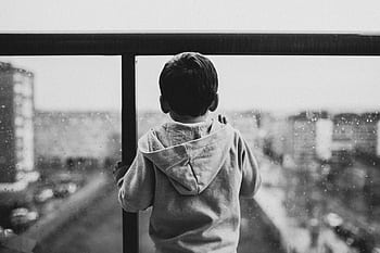 A young boy in a hoodie looks out a rainy window, creating a moody black-and-white scene. HD wallpaper