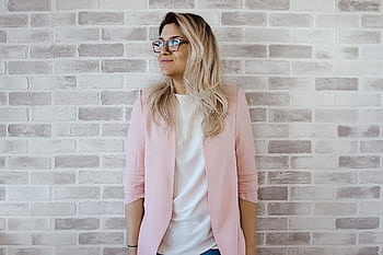 Elegant woman with glasses poses against a brick wall in a trendy pink blazer. HD wallpaper