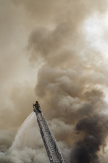 Brave firefighter extinguishing fire from a ladder amidst heavy smoke backdrop. HD phone wallpaper