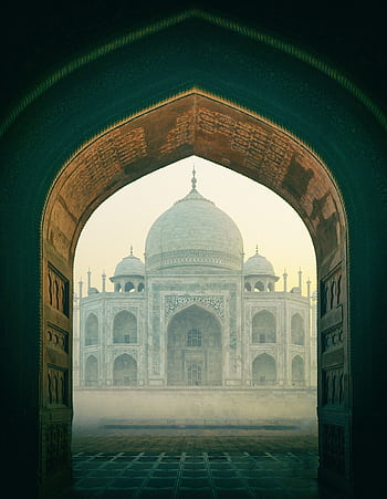 The iconic Taj Mahal viewed through a historic archway, enveloped in mist during sunrise in Agra, India. HD phone wallpaper