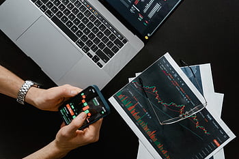 Hands using smart beside laptop with stock charts, showcasing digital trading. HD wallpaper