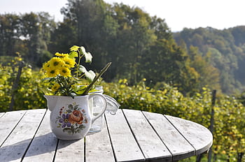 Yellow flowers in a decorative vase on a wooden table in a serene outdoor setting with trees. HD wallpaper