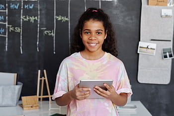 Cheerful girl holding a tablet in a modern classroom with a chalkboard background. HD wallpaper