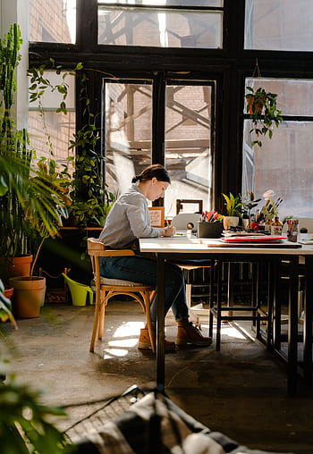 A female artist working diligently in a sunlit studio surrounded by plants and art supplies. HD phone wallpaper