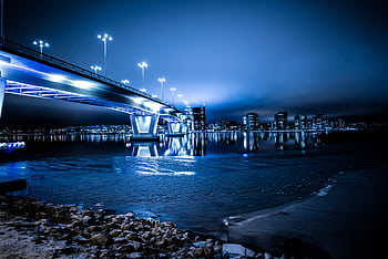 A vibrant blue cityscape with a modern illuminated bridge reflected in calm waters at night. HD wallpaper