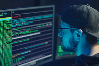 A cybersecurity expert inspecting lines of code on multiple monitors in a dimly lit office. HD wallpaper