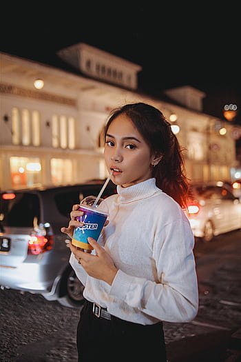 Stylish woman in a white sweater enjoys a drink on a bustling city street at night. HD phone wallpaper