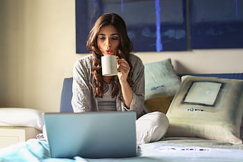 A woman enjoying coffee while working from home in a cozy bedroom setting with a laptop. HD wallpaper