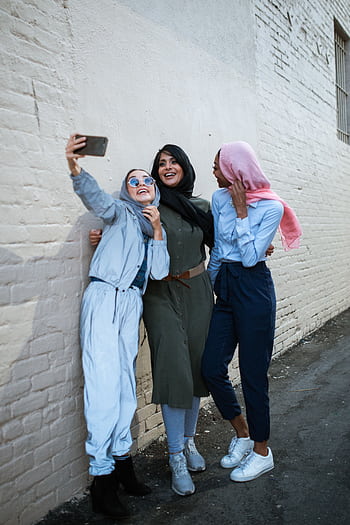 Three diverse friends in hijabs joyfully taking a selfie against a brick wall. HD phone wallpaper