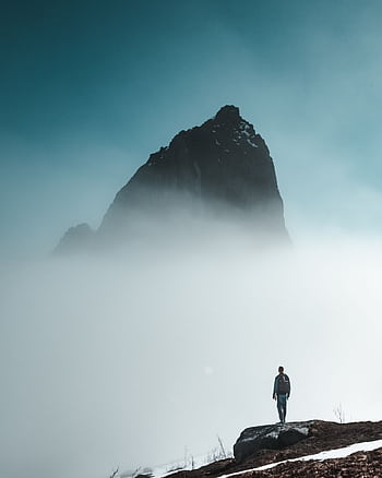 Man stands on rocky terrain surrounded by dense fog with towering mountain in background. HD phone wallpaper