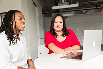 Two women engaging in a collaborative discussion in a modern office setting, using a laptop. HD wallpaper