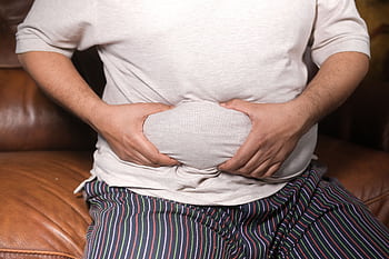 A close-up view of a man holding his tummy while sitting on a couch, highlighting lifestyle choices. HD wallpaper