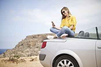 Smiling young woman sitting on a car at the beach in Mgarr, Malta using a smart. HD wallpaper