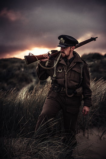 A soldier in uniform carries a rifle through grassy dunes at sunset. HD phone wallpaper
