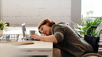 Woman in wheelchair sleeping at desk with laptop, illustrating exhaustion and remote work challenges. HD wallpaper