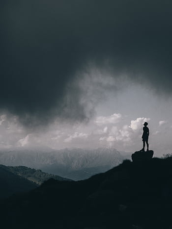 Silhouette of a lone person standing on a mountain peak under dramatic, dark clouds. HD phone wallpaper