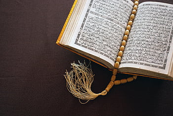 A Quran open to Arabic text with prayer beads resting on a brown surface, symbolizing Islamic faith. HD wallpaper