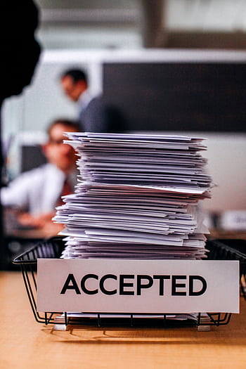 Pile of papers with 'Accepted' label in a busy office setting, highlighting information management. HD phone wallpaper