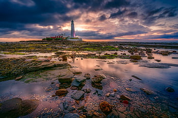 Dramatic sunset over St Mary's Lighthouse with reflections on mossy rocks and a cloudy sky. HD wallpaper