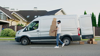 Man unloading cardboard boxes from a delivery van in a suburban neighborhood street. HD wallpaper