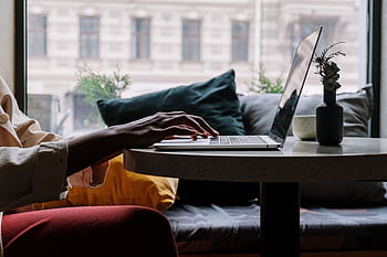 Close-up of a person typing on a laptop at a cozy cafe table with cushions. HD wallpaper