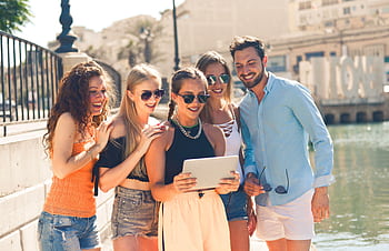 Five friends laughing together by the waterfront in Malta, enjoying a sunny day and sharing a tablet. HD wallpaper